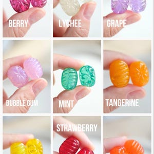 May include: A collection of colorful, jewel-toned candies in various flavors, including berry, lychee, grape, bubble gum, mint, tangerine, lemon, strawberry, and orange. Each candy is held between fingers, with the flavor name displayed below.