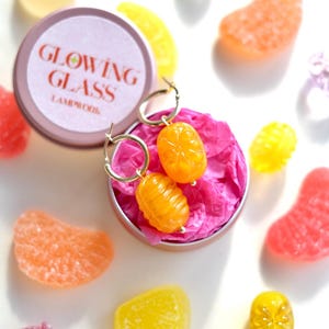 May include: Gold hoop earrings with orange, citrus-shaped charms rest in a small, pink-lined tin. The tin is surrounded by colorful, candy-like shapes in shades of orange, yellow, pink, and purple. The tin lid reads "GLOWING GLASS LAMPWORK".