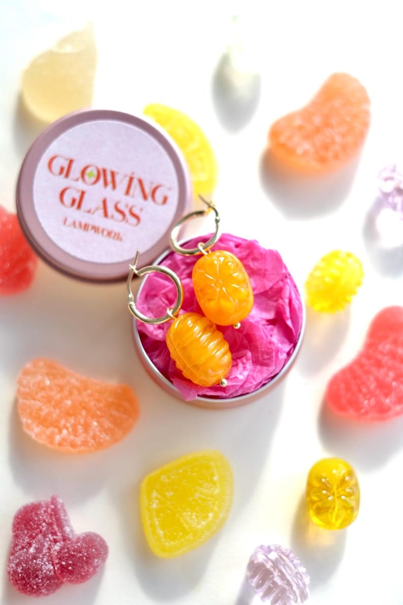 May include: Gold hoop earrings with orange, citrus-shaped charms rest in a small, pink-lined tin. The tin is surrounded by colorful, candy-like shapes in shades of orange, yellow, pink, and purple. The tin lid reads "GLOWING GLASS LAMPWORK".