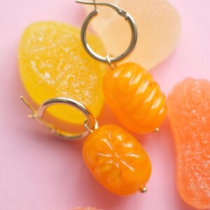 May include: Gold hoop earrings with orange, citrus-shaped charms. The earrings are set against a pink background with colorful gummy candies in shades of orange and yellow. The charms have a glossy finish.