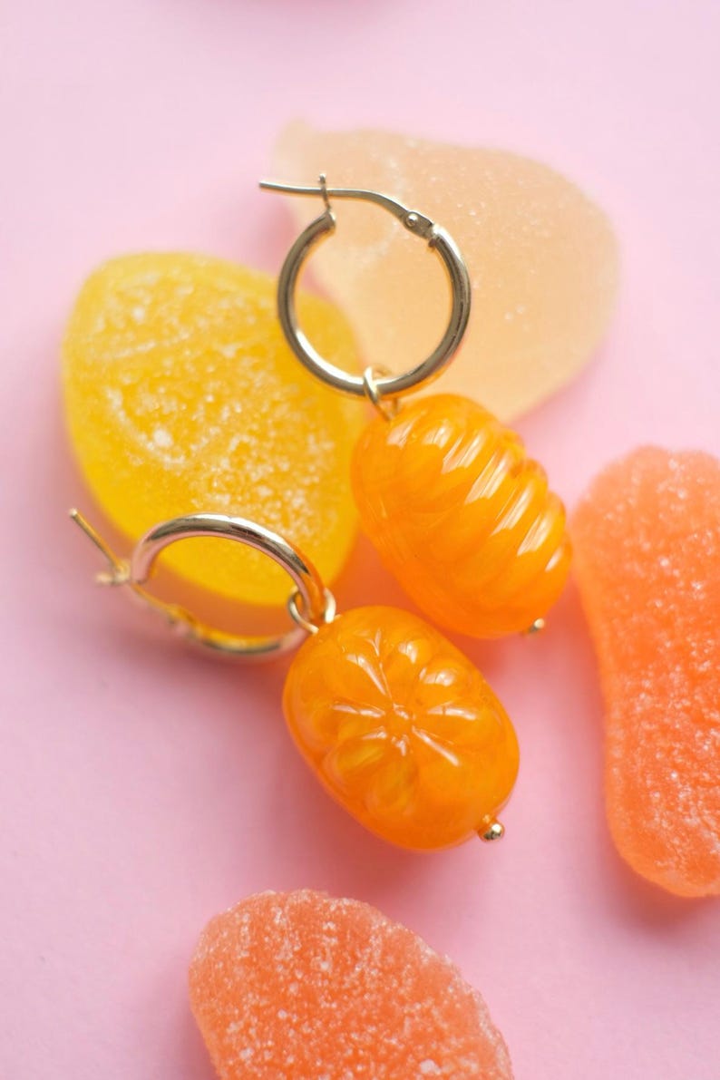 May include: Gold hoop earrings with orange, citrus-shaped charms. The earrings are set against a pink background with colorful gummy candies in shades of orange and yellow. The charms have a glossy finish.