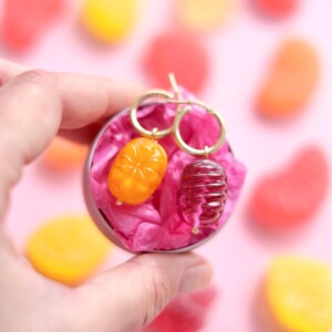 May include: A pair of gold hoop earrings with unique charms. One charm is orange and oval-shaped, the other is a dark red, cylindrical shape. The earrings are presented in a small, round tin filled with pink tissue paper, surrounded by colorful candies.