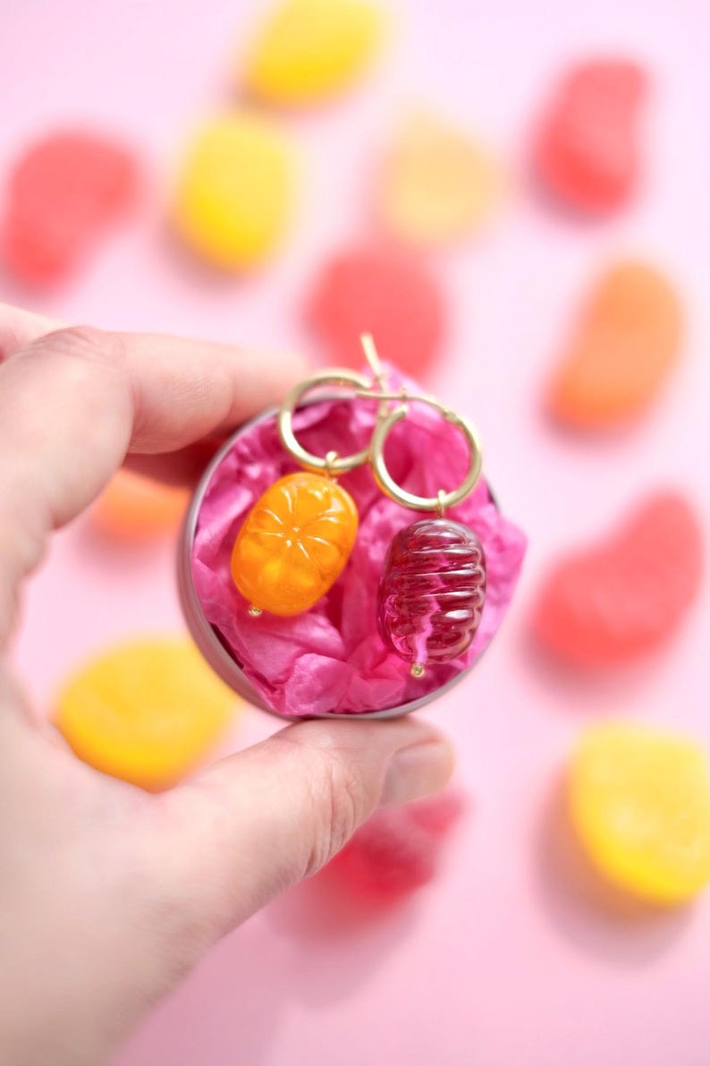 May include: A pair of gold hoop earrings with unique charms. One charm is orange and oval-shaped, the other is a dark red, cylindrical shape. The earrings are presented in a small, round tin filled with pink tissue paper, surrounded by colorful candies.