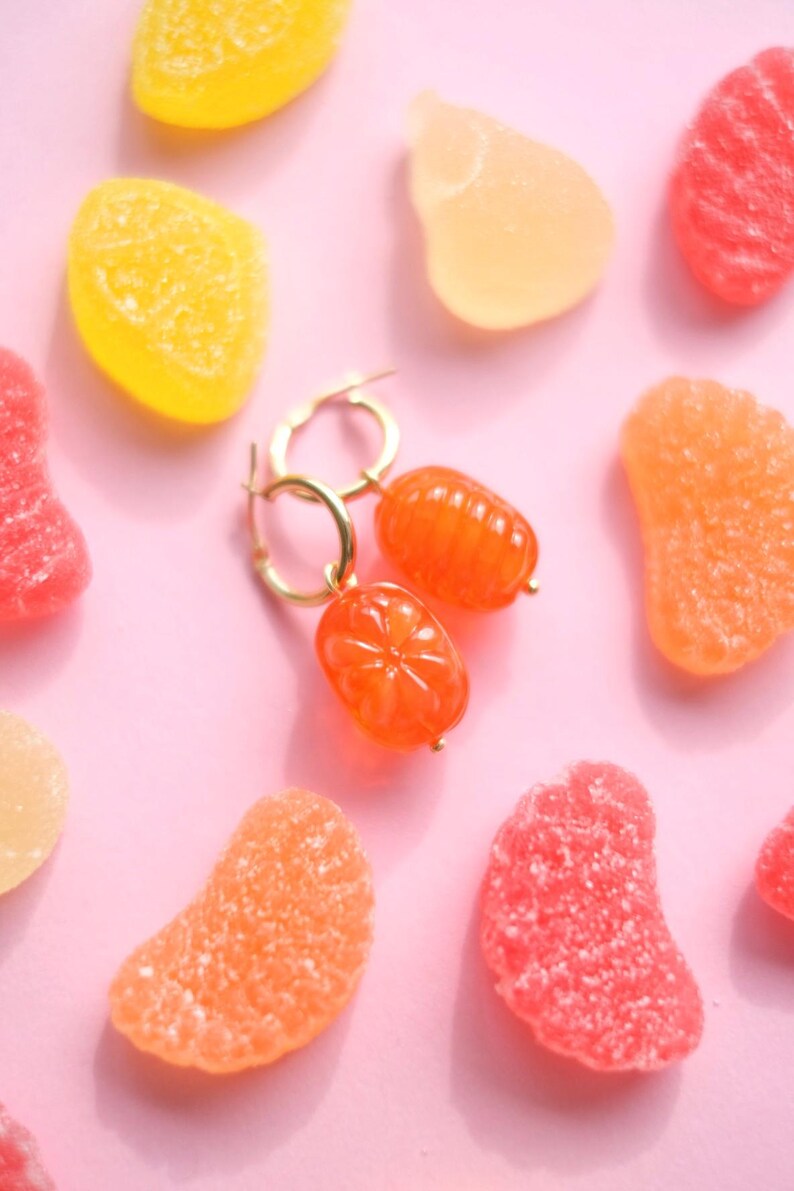 May include: Gold hoop earrings with orange, citrus-shaped charms. The earrings are displayed on a pink background scattered with colorful gummy candies in shades of orange, yellow, and red. A playful and vibrant image.