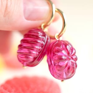 May include: A pair of pink, translucent earrings with gold-colored hoops. Each earring features a detailed, carved design. One earring has a ribbed texture, while the other has a floral pattern. The earrings are held up against a soft, blurred background.