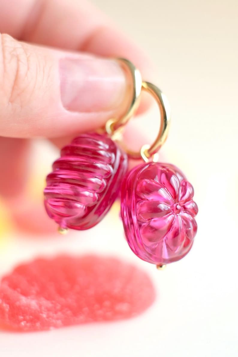 May include: A pair of pink, translucent earrings with gold-colored hoops. Each earring features a detailed, carved design. One earring has a ribbed texture, while the other has a floral pattern. The earrings are held up against a soft, blurred background.