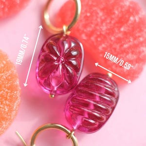 May include: A pair of pink, gold-toned earrings. The earrings are 19mm/0.74" and 15mm/0.59" in size, with a weight of about 6.5 grams each. The earrings have a gold hoop and a pink, oval-shaped, transparent charm.