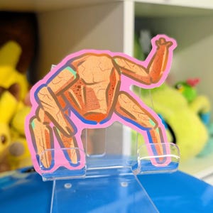 May include: A clear acrylic phone stand supporting a cartoon crab sticker. The crab is orange and tan with a pink outline and blue accents. The stand is on a blue surface, with a white shelf and soft toys in the background.