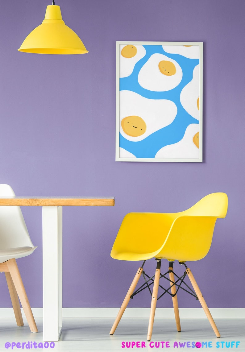 May include: A framed print featuring a blue background with a pattern of white eggs with yellow yolks and smiling faces. The print is hanging on a purple wall above a yellow chair and a white table with a wooden top.