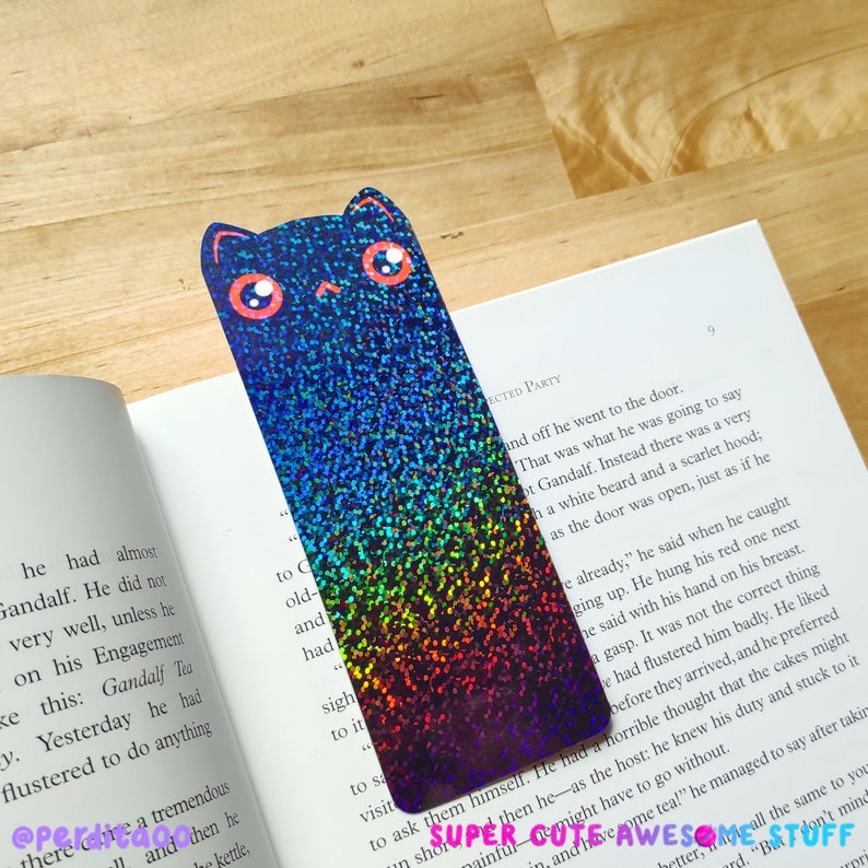 May include: A holographic rainbow cat bookmark with pink eyes. The bookmark is made of iridescent glitter paper and has a black outline.