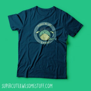 May include: A navy blue t-shirt featuring a cartoon dragon illustration inside a circular frame. The dragon is green with yellow stripes and wings. The text "Dragons are cute, It is known" is above and below the dragon. The website address is at the bottom.