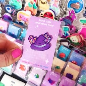 Könnte beinhalten: Ein lila Hexenhut mit Kristallen oben drauf. Der Hut ist eine handgefertigte Emaille-Anstecknadel mit glänzender Oberfläche. Die Anstecknadel ist in einer durchsichtigen Plastiktüte mit einer rosa und weißen Karte verpackt, auf der "Super Cute Handmade Pins!" und "Super Cute Awesome Stuff" steht.