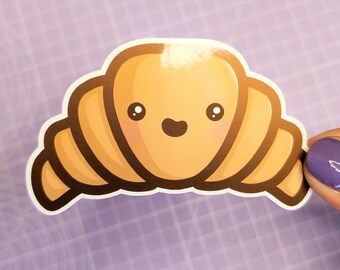 May include: A cartoon croissant sticker with a happy face. The sticker is brown and yellow and has a glossy finish.