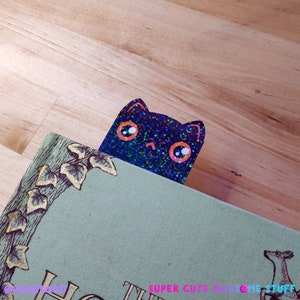 May include: A black cat bookmark with iridescent glitter and orange eyes peeking out from behind a book with green ivy and a deer illustration.