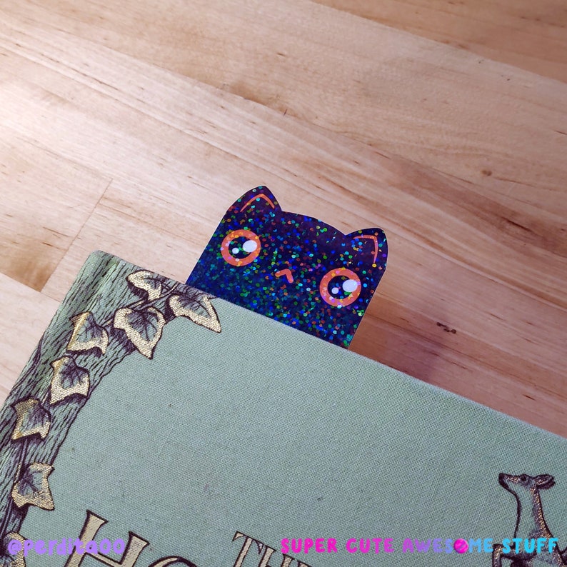 May include: A black cat bookmark with iridescent glitter and orange eyes peeking out from behind a book with green ivy and a deer illustration.