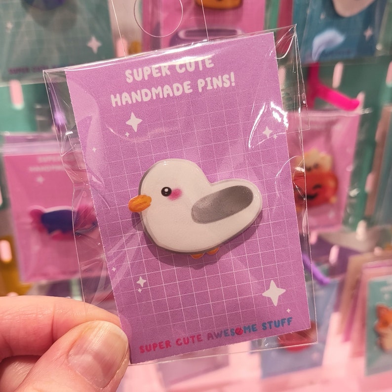 May include: A handmade pin featuring a cartoon seagull with a white body, gray wings, and an orange beak and feet. The pin is packaged on a purple card with the text "SUPER CUTE HANDMADE PINS!" and "SUPER CUTE AWESOME STUFF".