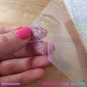 May include: A sheet of clear iridescent vinyl with a holographic finish. The vinyl is covered in small, iridescent dots. The vinyl is being held in a hand.