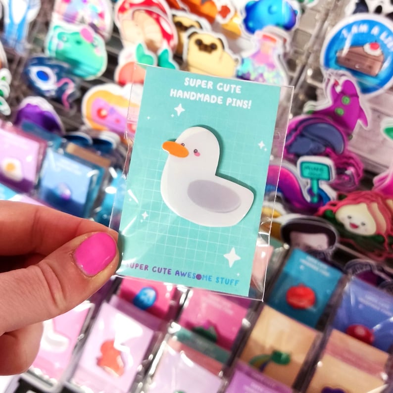 May include: A white duck pin with an orange beak and gray accents is displayed on a teal card. The card reads "SUPER CUTE HANDMADE PINS!" and "SUPER CUTE AWESOME STUFF". The pin is in a clear plastic sleeve, surrounded by other colorful pins.