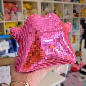 Op de afbeelding: Een roze, stervormig decoratief object bedekt met kleine, reflecterende vierkantjes. Het object heeft een textuur. Op de achtergrond zijn planken vol pluche speelgoed te zien. Het object wordt in een hand gehouden.