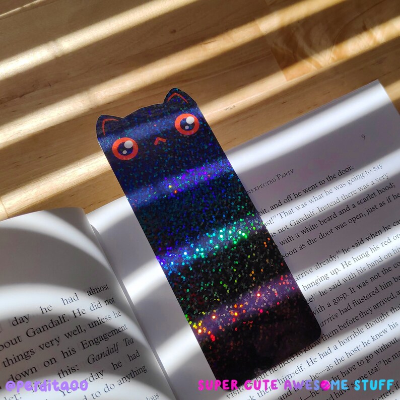 May include: A black cat bookmark with orange eyes and a glittery iridescent finish. The bookmark is shaped like a cat with a simple design. The text "SUPER CUTE AND AWESOME STUFF" is visible at the bottom of the image.