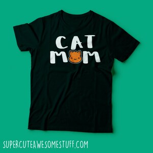 May include: Black t-shirt with the words "CAT MOM" in white block letters. The "O" in "MOM" is replaced with a cartoon cat face in shades of orange and brown. The website address "SUPERCUTEAWESOMESTUFF.COM" is printed at the bottom.