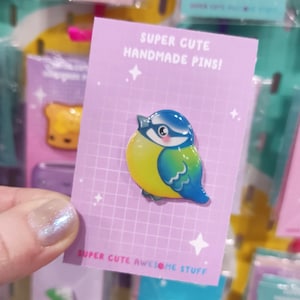 May include: A bluebird pin with a yellow belly and blue wings. The pin is on a purple card with white grid lines and the text "Super Cute Handmade Pins!" and "Super Cute Awesome Stuff".