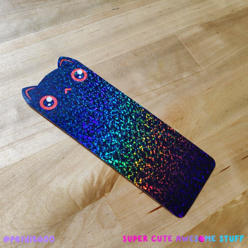 May include: A black cat bookmark with iridescent glitter and rainbow colors. The cat has large, round eyes with red pupils.