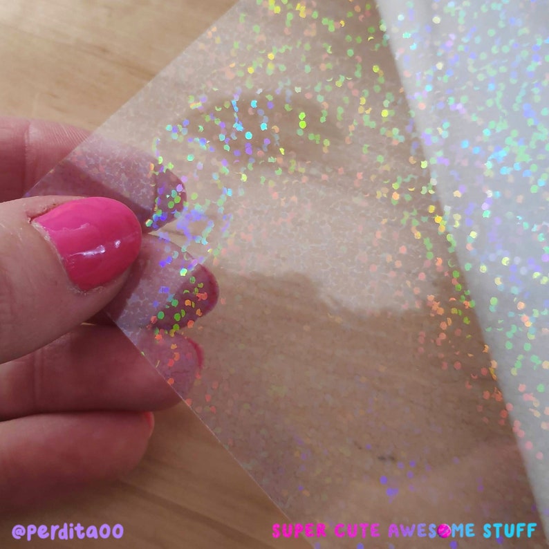 May include: A sheet of clear iridescent holographic vinyl with a scattered pattern of small, round, holographic dots. The vinyl is being held by a hand with pink nail polish.