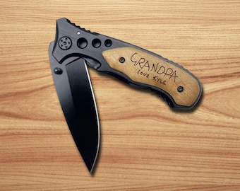 May include: A folding pocket knife with a matte black blade and a black metal frame featuring a light wood handle inlay. The wood handle is engraved with the text "GRANDPA love KYLE" in a handwritten style. The knife has a pivot screw and circular cutouts.