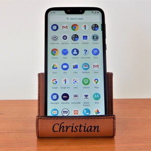 May include: A brown leather phone stand with the name "Christian" engraved on the front. A smartphone is in the stand, displaying a variety of apps on the screen.