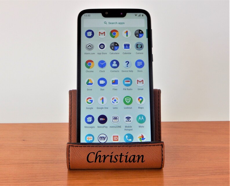 May include: A brown leather phone stand with the name "Christian" engraved on the front. A smartphone is in the stand, displaying a variety of apps on the screen.