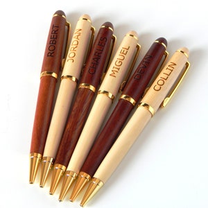 May include: A collection of personalized wooden pens. The pens feature gold-colored accents and are engraved with names such as "ROBERT", "JORDAN", and "CHARLES". The pens are made of light and dark wood.