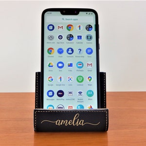 May include: A black leather phone stand with a gold inscription that reads "amelia". A smartphone is in the stand, displaying a screen full of app icons.