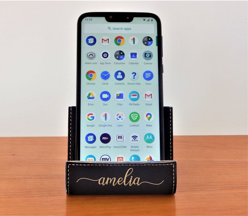 May include: A black leather phone stand with a gold inscription that reads "amelia". A smartphone is in the stand, displaying a screen full of app icons.