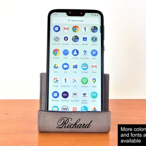 May include: A gray leather phone stand with the name "Richard" engraved on the front. The stand is holding a smartphone with a variety of apps displayed on the screen. The stand is on a wooden surface.