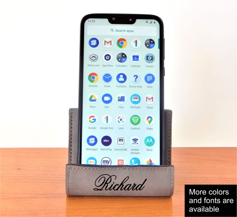 May include: A gray leather phone stand with the name "Richard" engraved on the front. The stand is holding a smartphone with a variety of apps displayed on the screen. The stand is on a wooden surface.