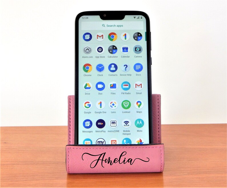 May include: A pink leather phone stand with the name "Amelia" engraved on the front. The stand is holding a black smartphone with a screen displaying a variety of apps.