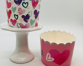 May include: Two cupcake liners with a scalloped edge. The liner on the left has a white background with a pattern of pink, purple, and blue hearts. The liner on the right has a pink background with a large pink heart and a smaller white heart.