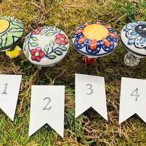 May include: Four ceramic mushroom-shaped ornaments with different painted designs. The ornaments are numbered 1 through 4. The designs include a yellow and green floral pattern, a red and green floral pattern, a blue and orange floral pattern, and a blue and white floral pattern.