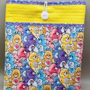 May include: A quilted fabric pouch featuring a colorful pattern of Care Bears. The pouch has a yellow top with white polka dots and a button closure. The main body of the pouch is covered in various Care Bear characters in shades of pink, blue, yellow, and purple.