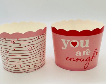 May include: Two pink and red cupcake liners. One liner has a white background with red stripes and small hearts. The other liner has a pink background with the text "you are enough" in white with a red heart.