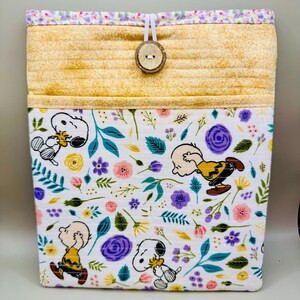 May include: A fabric storage container with a Snoopy and Charlie Brown floral design. The top section is yellow with horizontal stitching, and the top edge has a floral pattern. A wooden button and elastic loop secure the container.