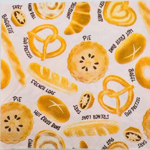 May include: A white napkin with illustrations of various baked goods, including croissants, pretzels, pies, and baguettes. The names of the pastries are printed in black text. The overall design is a collection of golden-brown baked items.