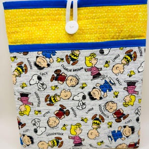 May include: A quilted fabric pouch featuring a gray background with a repeating pattern of Peanuts characters including Snoopy, Charlie Brown, Lucy, Linus, Sally, and Woodstock. The top section is yellow with white dots and a white button closure.