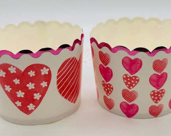 May include: Two white cupcake liners with pink scalloped edges. One liner has a large red heart with white polka dots and a smaller red heart with white stripes. The other liner has a vertical row of five red hearts with different patterns.