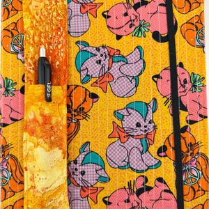 May include: A fabric notebook cover featuring a yellow background with a repeating pattern of pink and teal cartoon cats and peace signs. The cover includes an attached orange marbled pen holder containing a black S.GEL pen and a black elastic band.