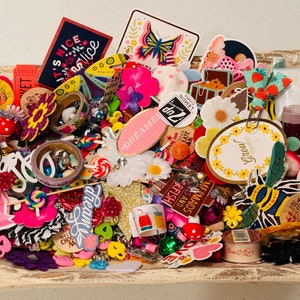 May include: A collection of colorful craft supplies, including stickers, washi tape, buttons, and charms. The supplies are arranged in a wooden bowl and feature a variety of themes, including flowers, butterflies, and animals.