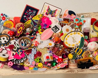 May include: A collection of colorful craft supplies, including stickers, washi tape, buttons, and charms. The supplies are arranged in a wooden bowl and feature a variety of themes, including flowers, butterflies, and animals.