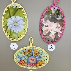 May include: Three oval-shaped crocheted ornaments with different floral images. The first ornament is yellow with a blue flower, the second is pink with pink flowers, and the third is yellow with a blue watering can and sunflowers.