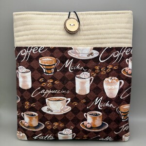 May include: A quilted book sleeve with a coffee-themed pattern. The brown fabric features coffee cups, beans, and the words "Coffee," "Mocha," "Cappuccino," and "Latte." It has a beige top with a button closure.